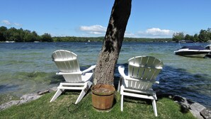 Two white Adirondack chairs by a tree on a grassy lakeside with clear water and boats in the background.