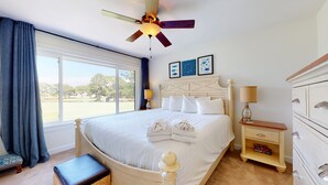Ein Hotelzimmer mit einem großen Bett, einem Deckenventilator, einem Nachttisch mit Lampe, einem Schminktisch und Blick ins Freie.