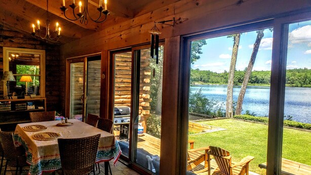 Interior de una cabaña de madera con comedor, una parrilla exterior y vista a un lago.