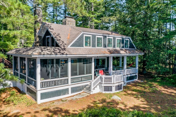 A cabin with a stone chimney, large windows, and a covered porch with steps leading up to it.