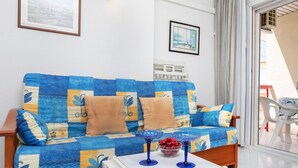 A living room with a blue and yellow patterned sofa, a white coffee table with a bowl of fruit, and a window with a view of the outside. - Apartment Ancora by Interhome (Salou)