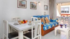 A dining area with a white table, chairs, and a blue and yellow sofa. There are framed pictures on the wall and an air conditioning unit above the table. - Apartment Ancora by Interhome (Salou)