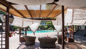 Un espacio de estar al aire libre techado con mobiliario de mimbre, una piscina al fondo y una estructura de pérgola.