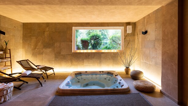 Un cuarto de spa con jacuzzi, sillones de descanso, un ventanal con vistas a la vegetación y una pared de piedra.