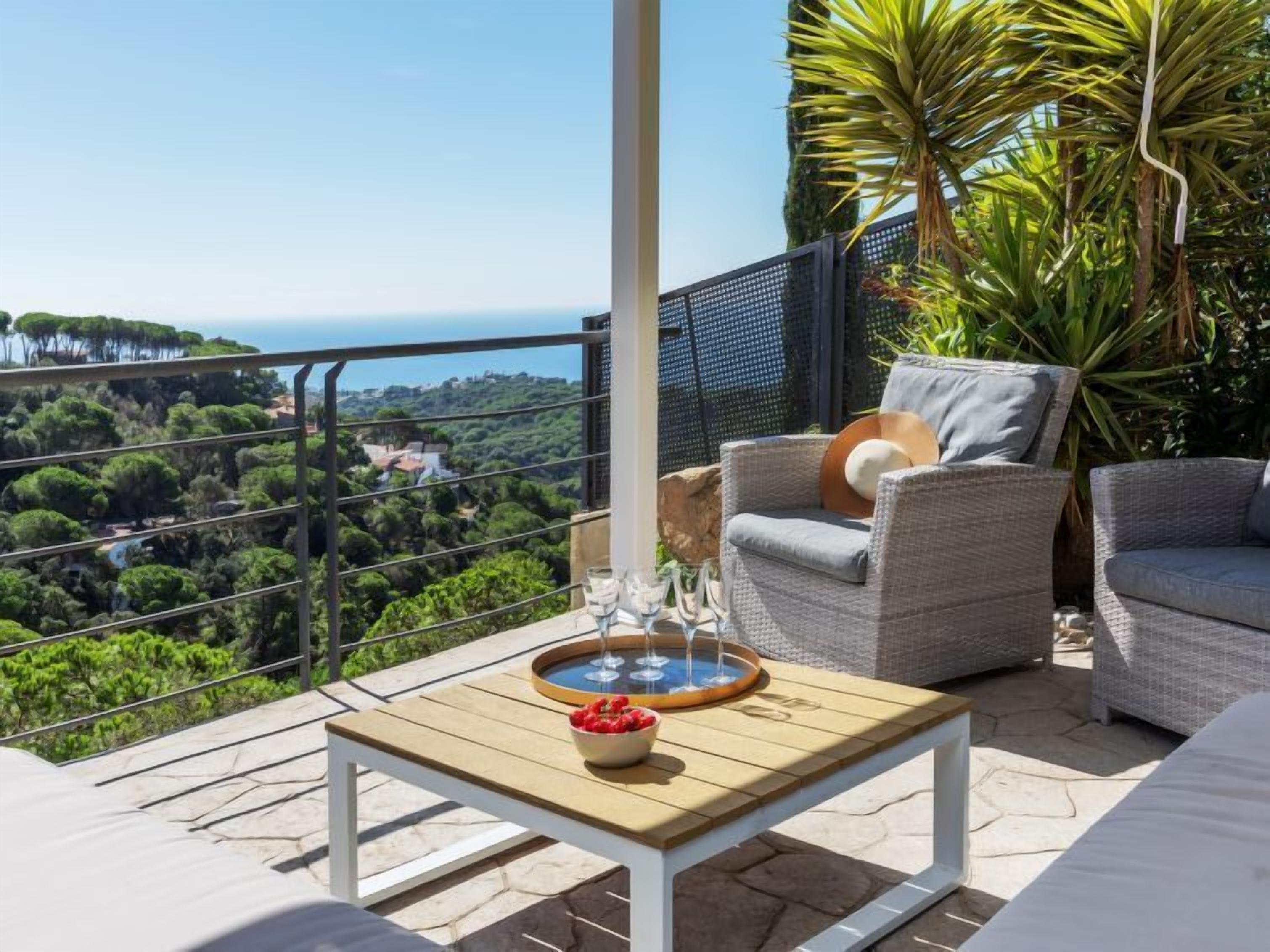 A balcony with wicker furniture, a table with a bowl of fruit, and a view of the sea and greenery.