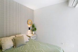 Un dormitorio con cama, almohadas, una mesita de noche con lámpara, un espejo en la pared y una planta en maceta.