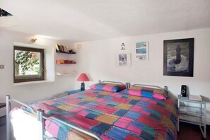 A bedroom with a metal bed frame, a colorful quilt, a window with a view of trees, a bookshelf, a wall-mounted lamp, and a framed picture.
