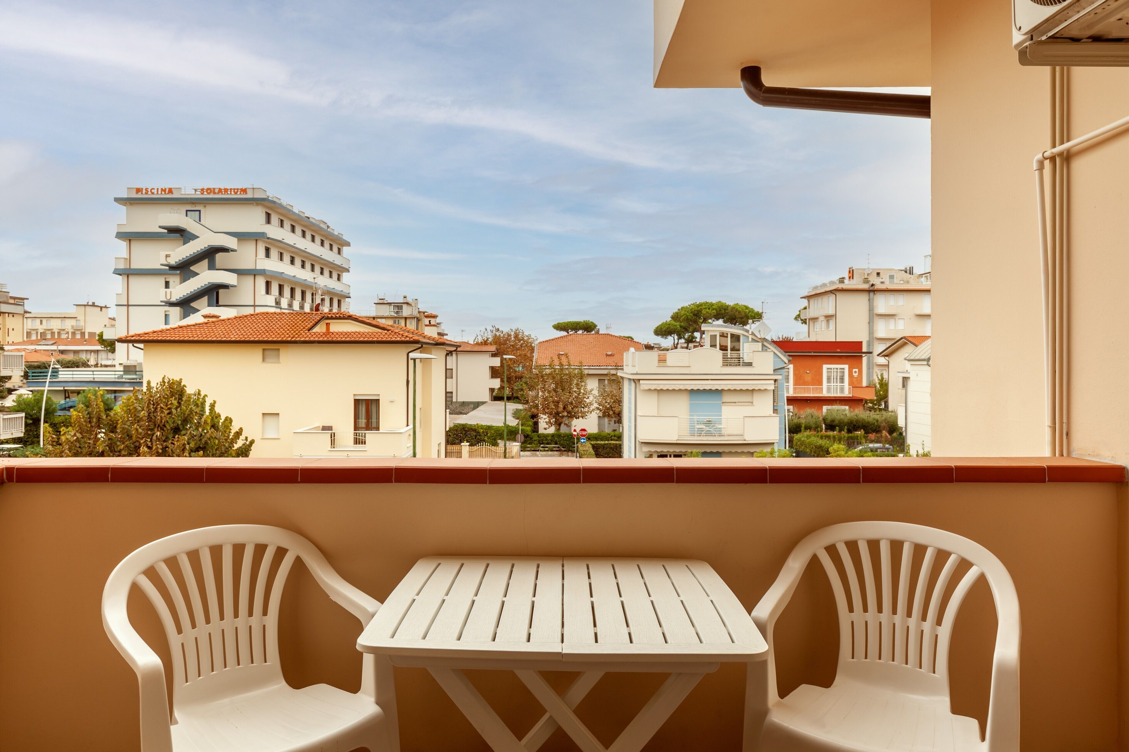 A balcony with two white plastic chairs and a small table, overlooking a residential area with buildings and trees.