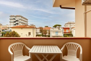 Un balcón con dos sillas de plástico blanco y una mesa pequeña, con vista a una zona residencial con edificios y árboles.