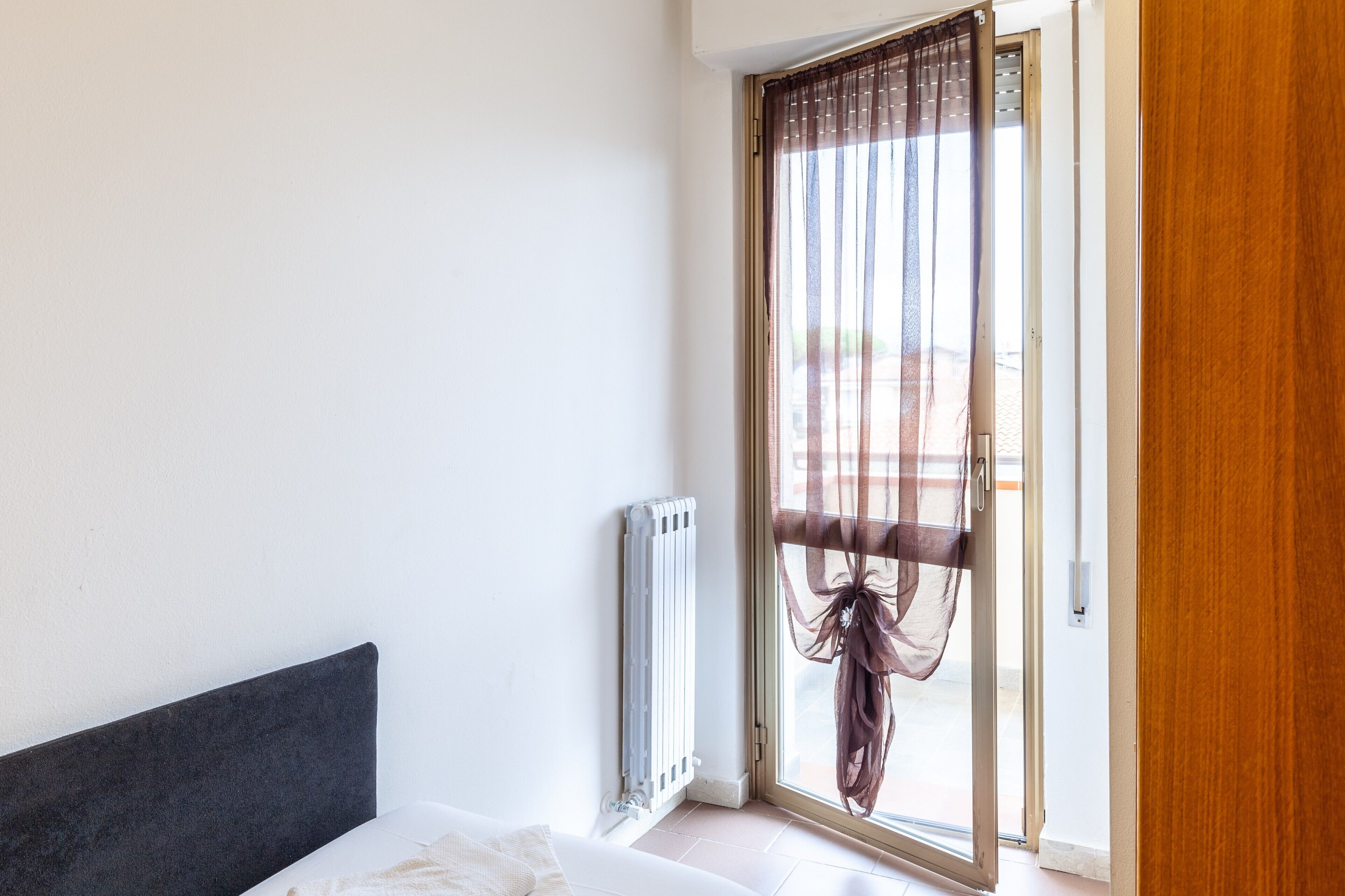 Quarto com cama, radiador e porta de vidro deslizante com cortinas leves.