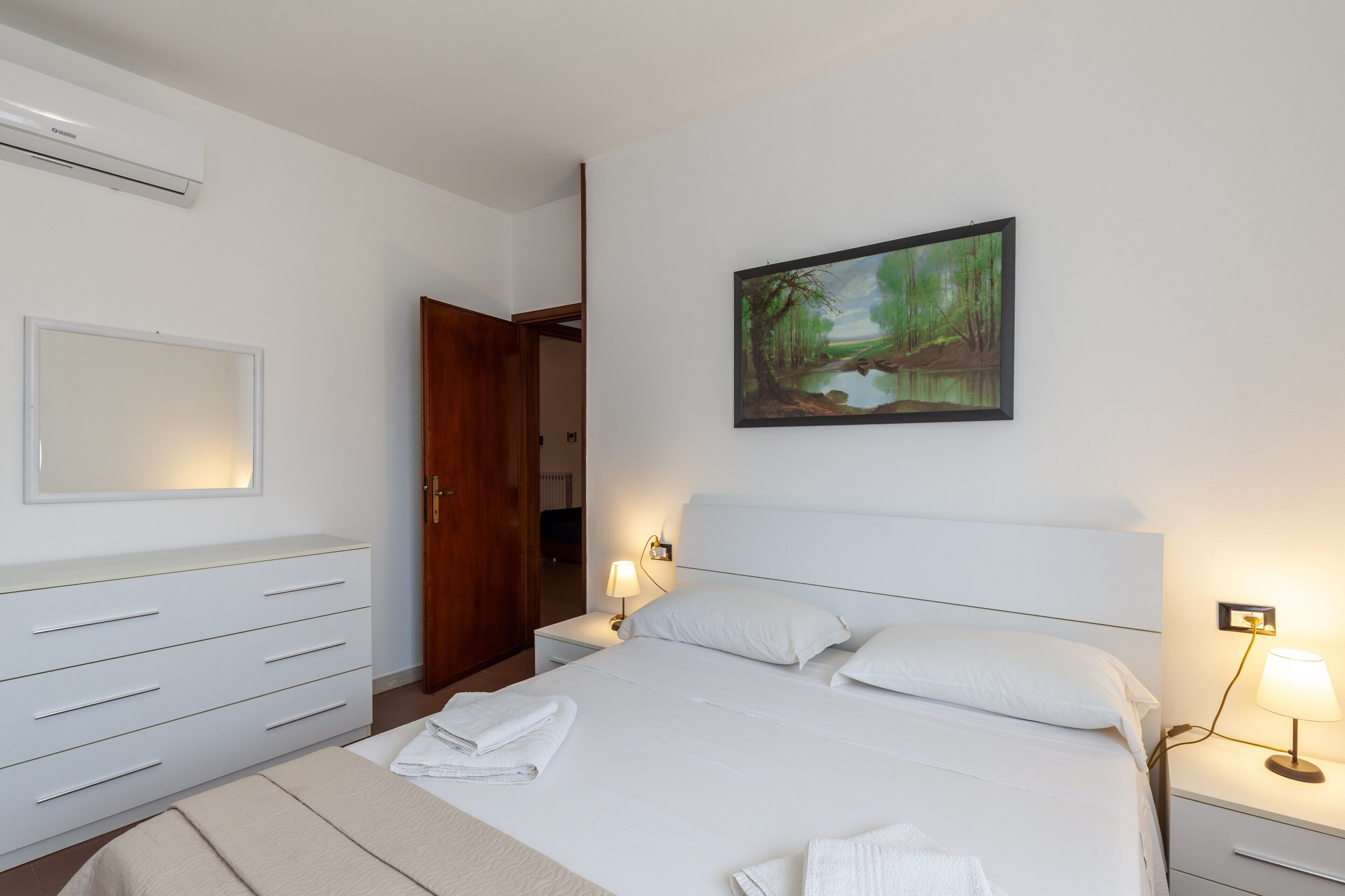A hotel room with a bed, bedside tables, a wardrobe, a painting, and an air conditioning unit.