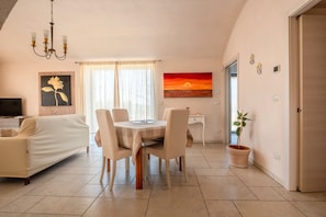 A dining area with a round table, chairs, a television, a sofa, a painting, a sunset view, a potted plant, and a door.