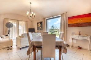 A modern living room with a dining area, a flat-screen TV, a chandelier, and a painting of a sunset.