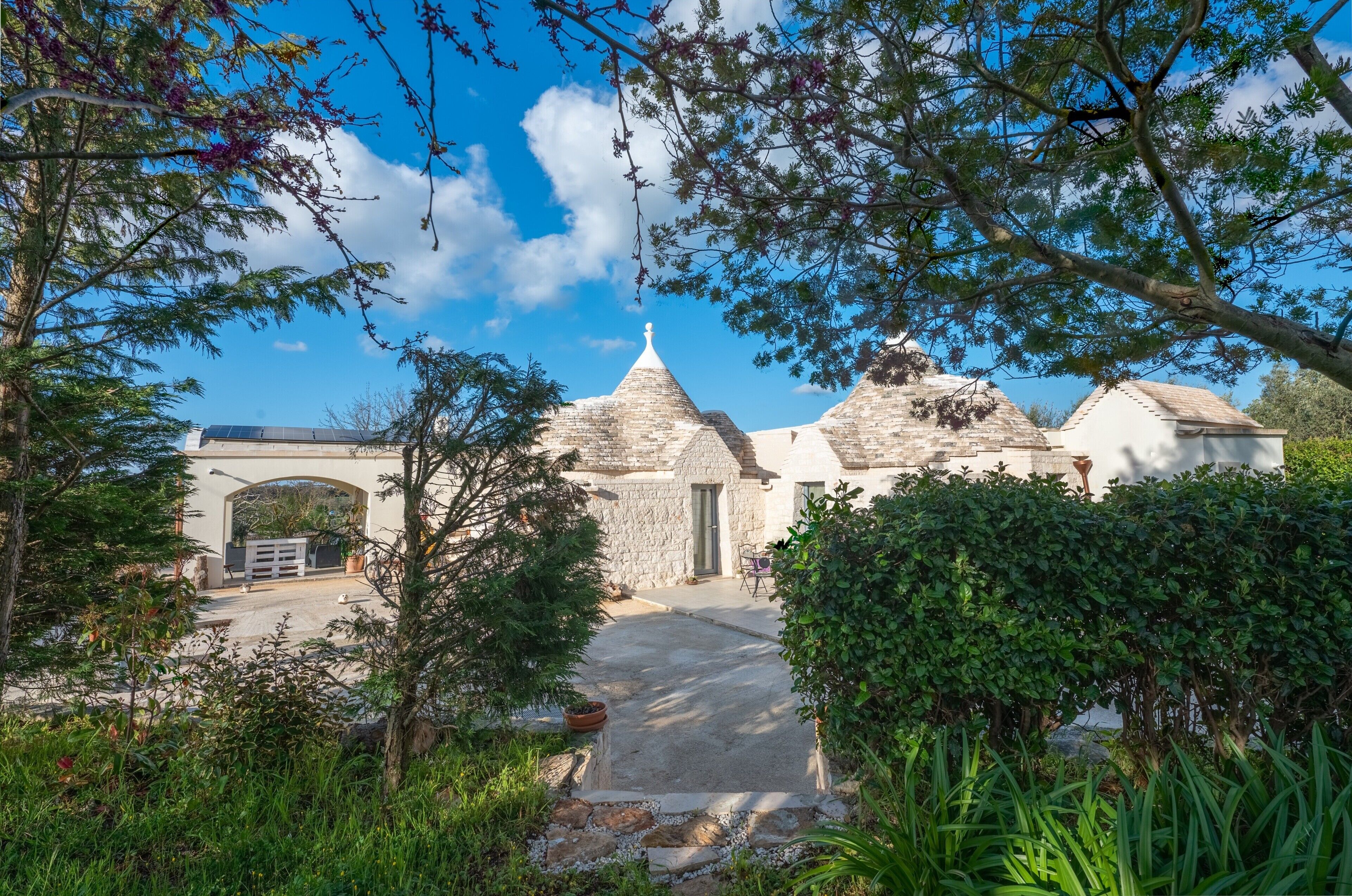 A stone building with a central dome and two smaller domes, surrounded by greenery and a clear sky.