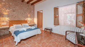A bedroom with a stone wall, wooden beams, a bed with white linens, a wooden bench, and a window with sheer curtains. - Finca "Can Miquel des Revellar" with Mountain View, Pool & Wi-Fi (Campos)