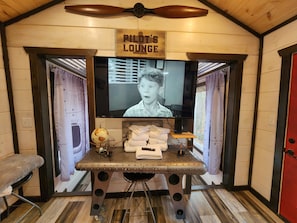 A room with a television mounted on a wall, a wooden sign above it reading "Pilot's Lounge," and a table with towels and a globe.