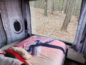 A bed with a colorful bedspread, a window with a view of a forest, and a metal wall with circular cutouts.