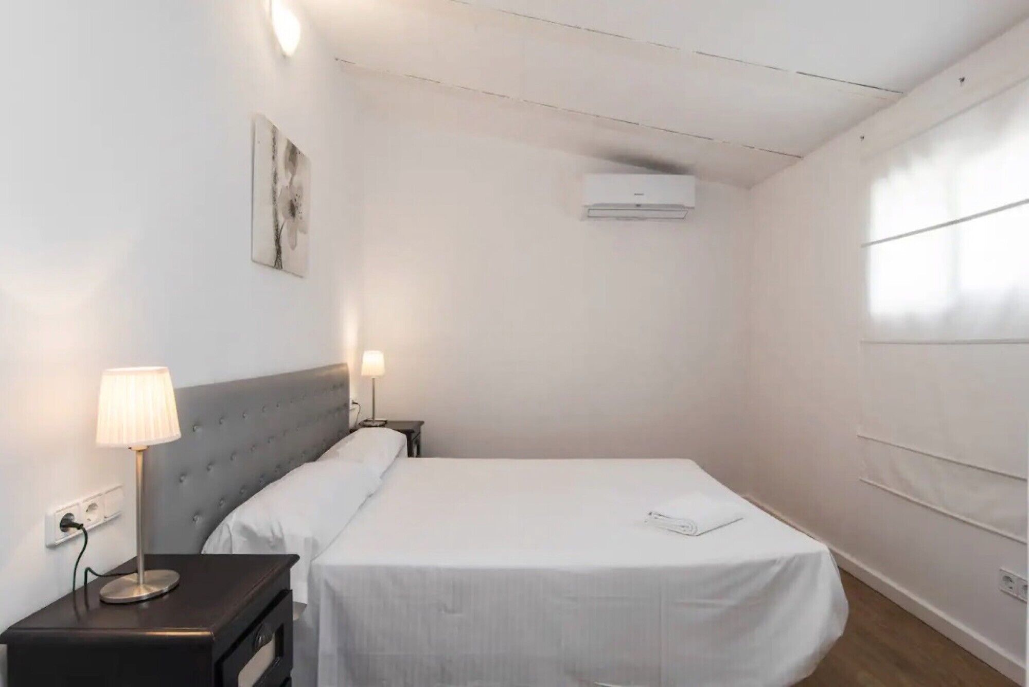 Une petite chambre à coucher, bien rangée, avec un lit simple, des lampes de chevet, un climatiseur fixé au mur et une fenêtre avec des stores.