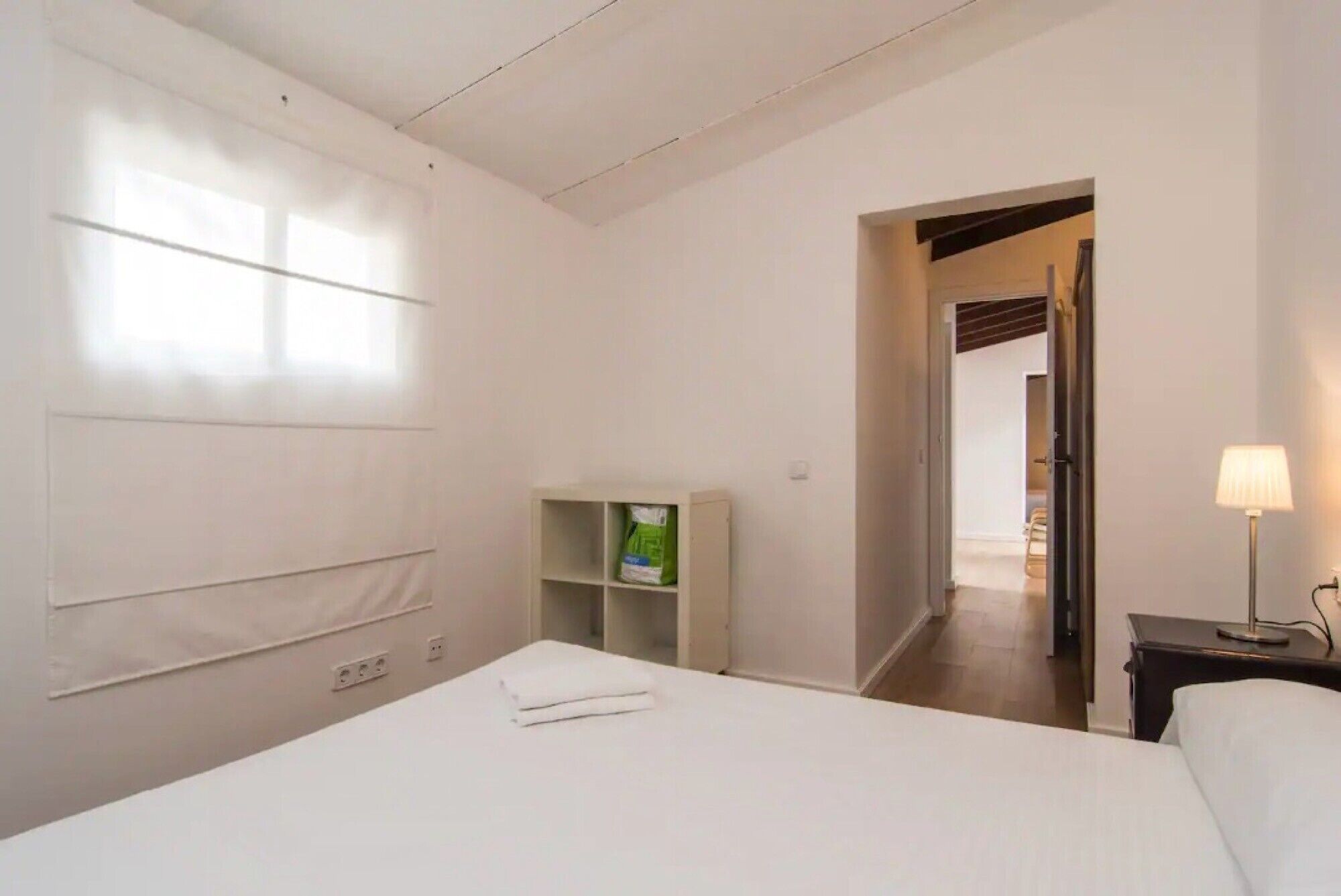 Una habitación de hotel con una cama, una mesita de noche con lámpara, un estante con maletas y una puerta que da a otra habitación.