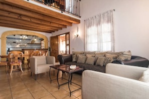 A living room with a sofa, chairs, a table, a kitchen in the background, and a balcony with a railing.