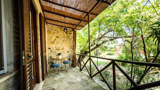 Un portico con pavimento in pietra, un tavolo e sedie, una porta in legno e una ringhiera metallica che si affaccia su un giardino.