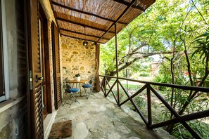 Un porche con suelo de piedra, una mesa, sillas, una puerta de madera y una barandilla metálica con vistas a un jardín.