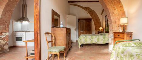 A room with a kitchen, two beds, a wooden chair, and a lamp.