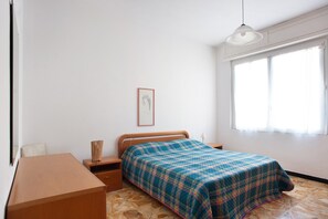 A bedroom with a bed, bedside tables, a desk, a chair, a window with curtains, and a hanging light fixture.