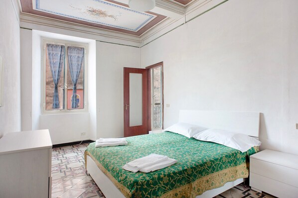 A bedroom with a bed, a desk, a window with curtains, a door, and a patterned floor.