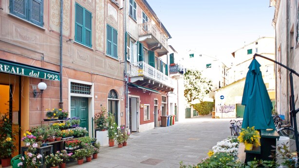 Un carrer estret amb edificis, terrasses a l'aire lliure i plantes en testos.