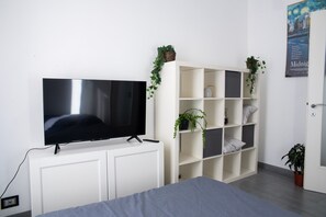 Un dormitorio con un televisor sobre un mueble blanco, una cama con ropa de cama gris, una estantería blanca con plantas y una puerta con un cartel.