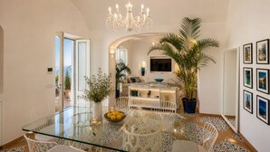 A modern dining area with a glass table, white chairs, a chandelier, and a large potted plant. - AMORE RENTALS - Villa Mare Blu with Internal Heated Pool, Sea View and Breakfast (Praiano)