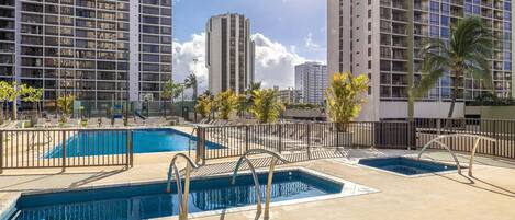 A rooftop pool area with multiple swimming pools, surrounded by tall buildings and palm trees.