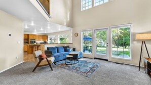 Un ampio soggiorno con soffitto alto, cucina integrata e grandi porte scorrevoli in vetro che si aprono su un giardino.
