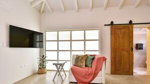 Ein modernes Wohnzimmer mit großem Fenster, Flachbildfernseher, einer Couch mit Decke, einem kleinen Tisch und einer Topfpflanze.