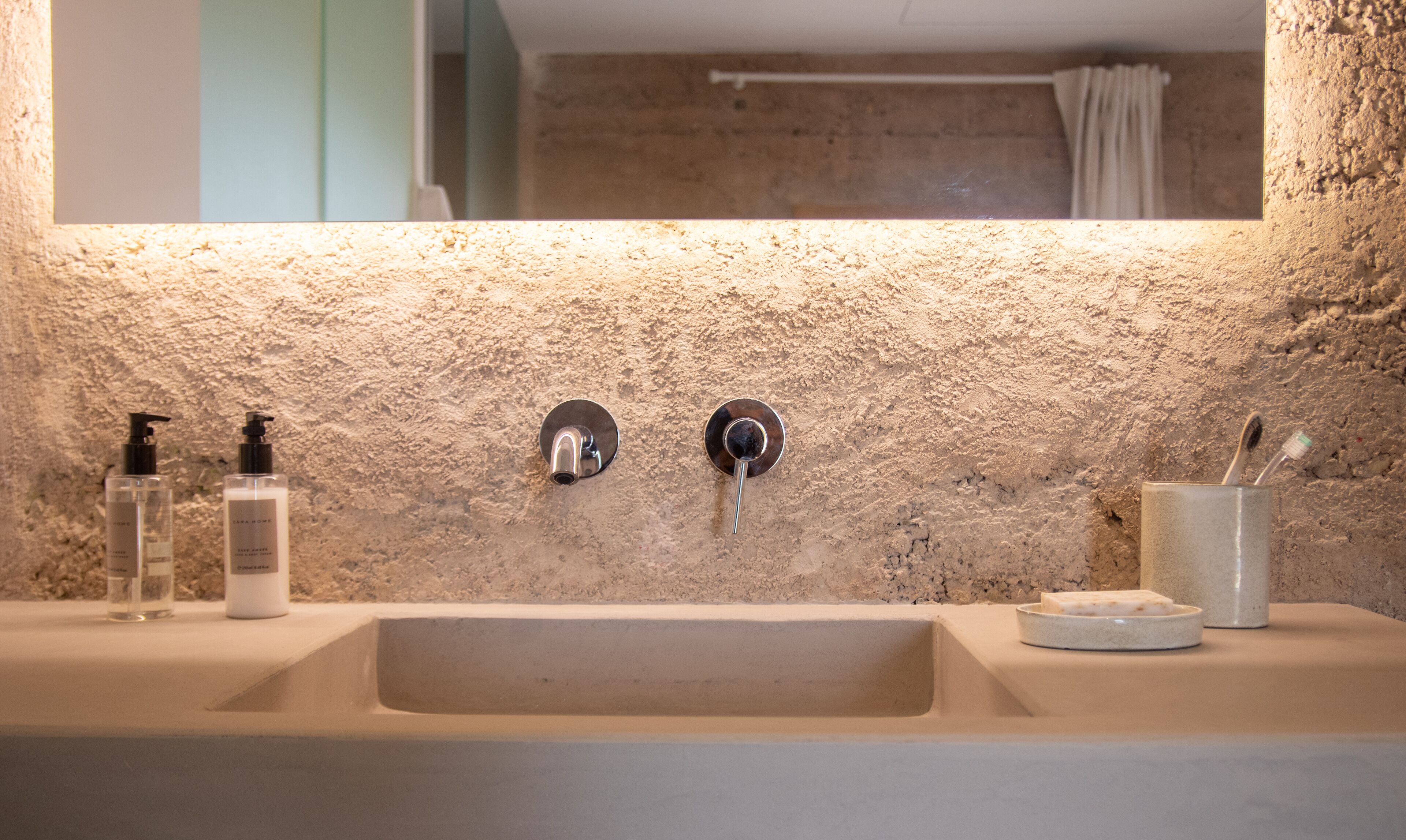 Une salle de bain avec un mur à texture, un lavabo, deux distributeurs de savon, un porte-brosse à dents et un miroir.