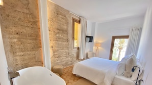 A modern bedroom with a large bathtub, a bed with white linens, a bedside table with a lamp, and a view of palm trees through a window. - Hotel Boutique Can Pocovi (Sant Llorenc des Cardassar)