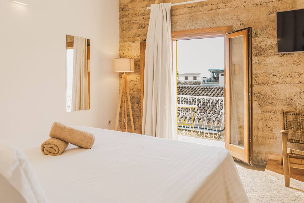 Een slaapkamer met een groot bed, een vloerlamp, een raam met gordijnen en uitzicht op gebouwen.