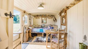 A cozy, rustic cabin interior with a wooden ceiling, a bed with a quilt, a dining table with chairs, and a hanging light fixture.