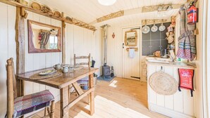 A rustic interior with a wooden dining table, chairs, a mirror, and a kitchen area with a stove and utensils.