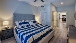 A bedroom with a large bed, a ceiling fan, a nightstand, and a door leading to a kitchen. - Crimson Court - Book Now for Football! (Tuscaloosa)