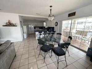A spacious living area with a dining table and chairs, a kitchen in the background, and a large window offering a view of the outside.