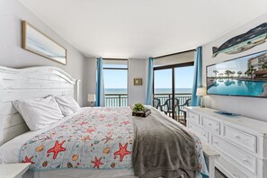 Ein Schlafzimmer mit einem Bett, einer Kommode, einem kleinen Tisch mit einer Pflanze und einem Bild von einer Strandansicht.
