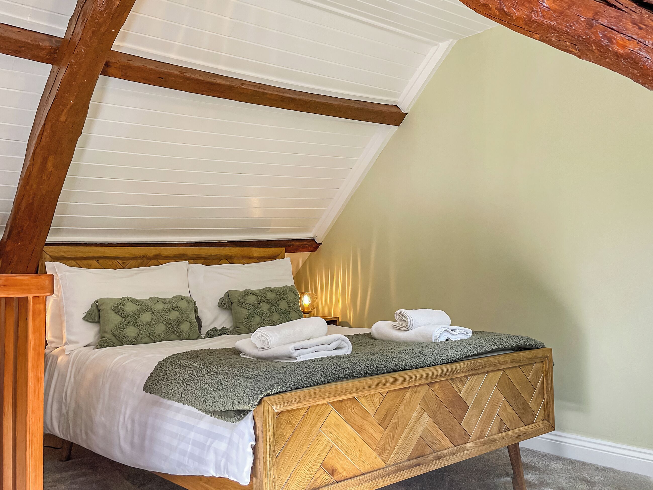 Een net opgemaakt bed met een houten hoofdbord, witte lakens en een groene plaids.