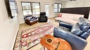 En stue med sofa, lænestol, sofabord, en guitar, et mønstret tæppe og et fjernsyn monteret på væggen.
