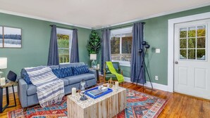 A living room with a grey sofa, a coffee table, and a green chair. There are two windows with curtains, a door, and a plant on the wall.