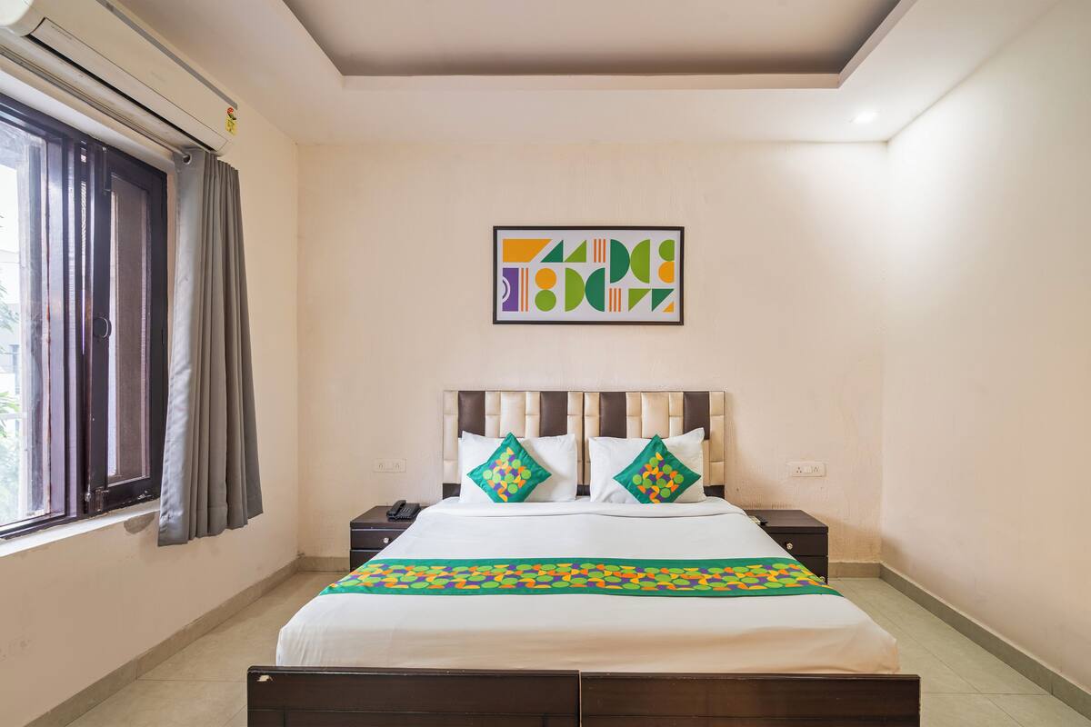 A hotel room with a bed, a nightstand, a wall-mounted air conditioner, a window with curtains, and a geometric wall art piece.