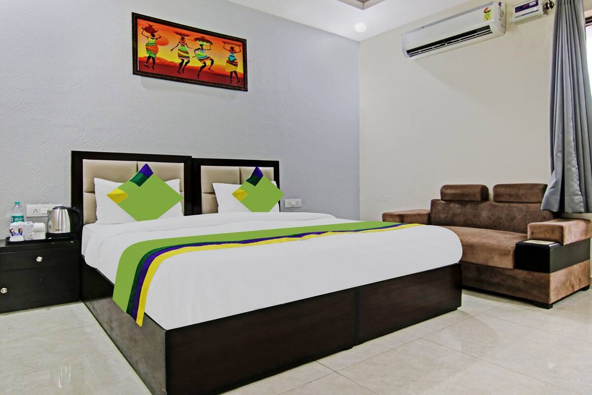 A hotel room with a bed, a nightstand, a coffee maker, a water bottle, a painting, a couch, and an air conditioning unit.