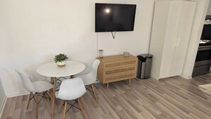 A small dining area with a round table, two chairs, a potted plant, a wooden chest of drawers, a trash bin, and a flat-screen TV mounted on the wall. - Sojourn | Bayside | 1 Bedroom (Norfolk)