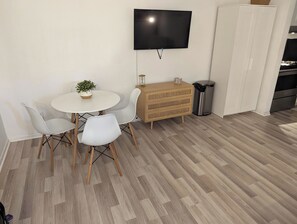 Un coin repas avec une table ronde, deux chaises blanches, une petite plante en pot, une commode en bois, une poubelle et un téléviseur fixé au mur.